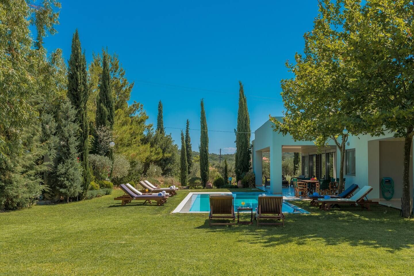 Zona junto a la piscina con sillas de descanso y una piscina, rodeada de árboles y un edificio con ventanas amplias.