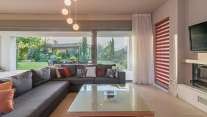 Un salon moderne avec un canapé d’angle, une table basse en verre et une grande fenêtre donnant sur un jardin.