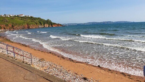 En kustnära strand med en strand av småsten, ett metallräcke och en fri utsikt över havet och de avlägsna kullarna.