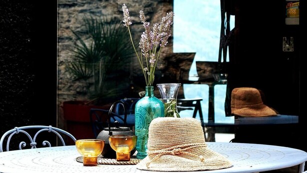 Una mesa redonda con un sombrero tejido, un florero de vidrio con flores y dos candelabros.