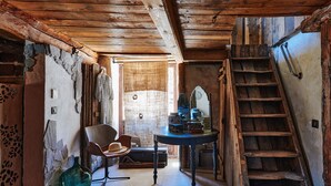 Una habitación rústica con una escalera de madera, una silla, una mesa con libros y una maleta antigua.