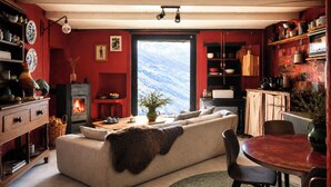 Una sala de estar acogedora con chimenea, un sofá, una mesa de comedor y una zona de cocina con paredes rojas y estantes de madera.