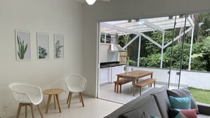 Ett modernt vardagsrum med en grå soffa, vita stolar, ett träbord och en stor glasdörr som leder ut till en balkong med utsikt över grönområden.