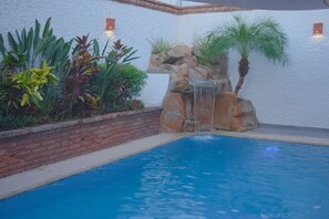 A swimming pool with a waterfall feature, surrounded by various plants and a brick wall. - Private house, pool, bbq and jacuzzi!! (Mazatlán)