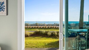 A view from a room with a glass door to a patio area with a view of the beach, a white fence, and outdoor furniture.