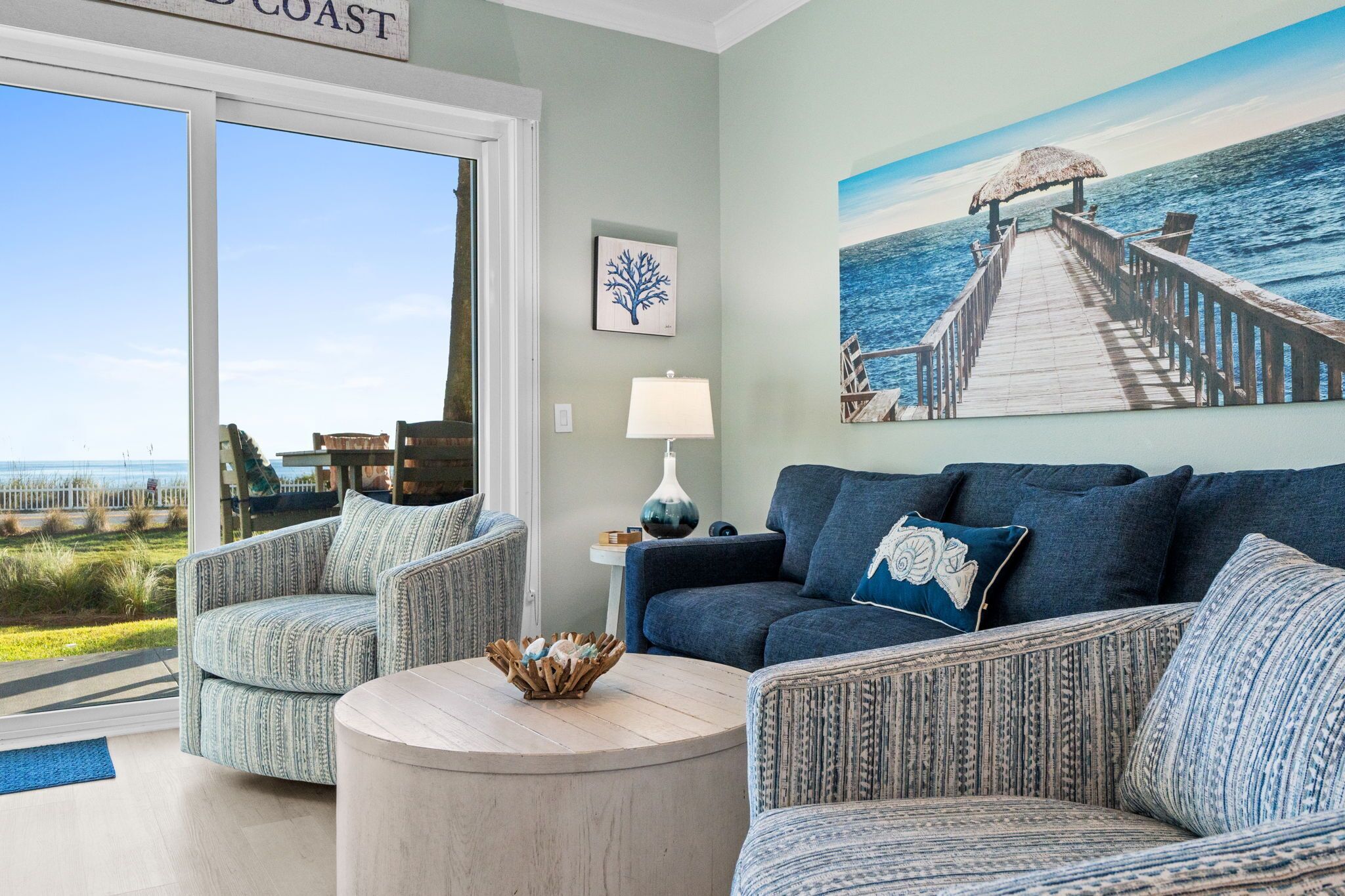 A living room with a view of the ocean, a wooden deck, and a painting of a pier.