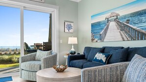 A living room with a view of the ocean, a wooden deck, and a painting of a pier.
