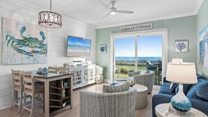 A spacious living room with a view of the Emerald Coast, featuring a dining area, comfortable seating, and a ceiling fan.