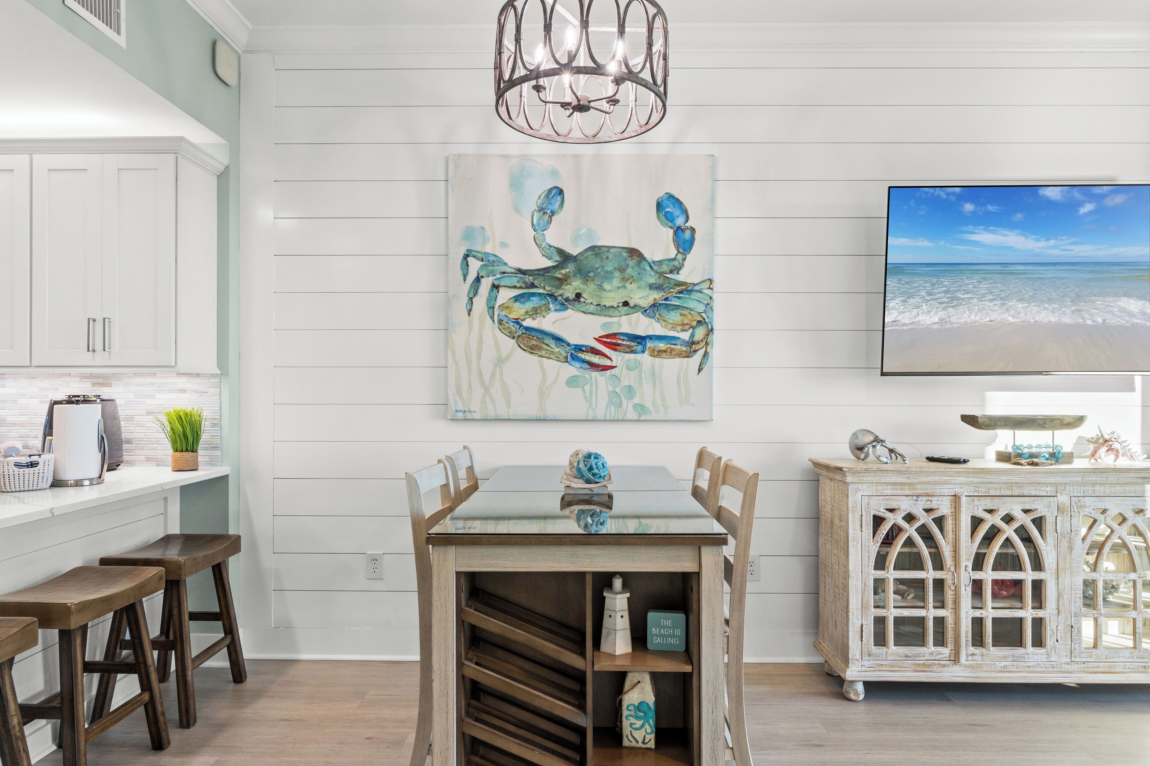 A kitchen with white cabinets, a wooden table with chairs, a painting of a crab, and a TV on the wall.