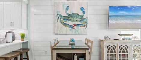 A kitchen with white cabinets, a wooden table with chairs, a painting of a crab, and a TV on the wall.