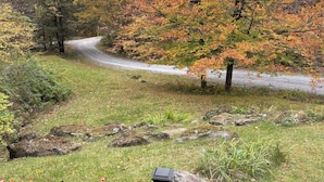 A wooden deck with a railing overlooking a grassy area with autumn-colored trees and a gravel path. - Canary Lodge - Berkshires Mountain Retreat - 6 Guests (West Stockbridge)