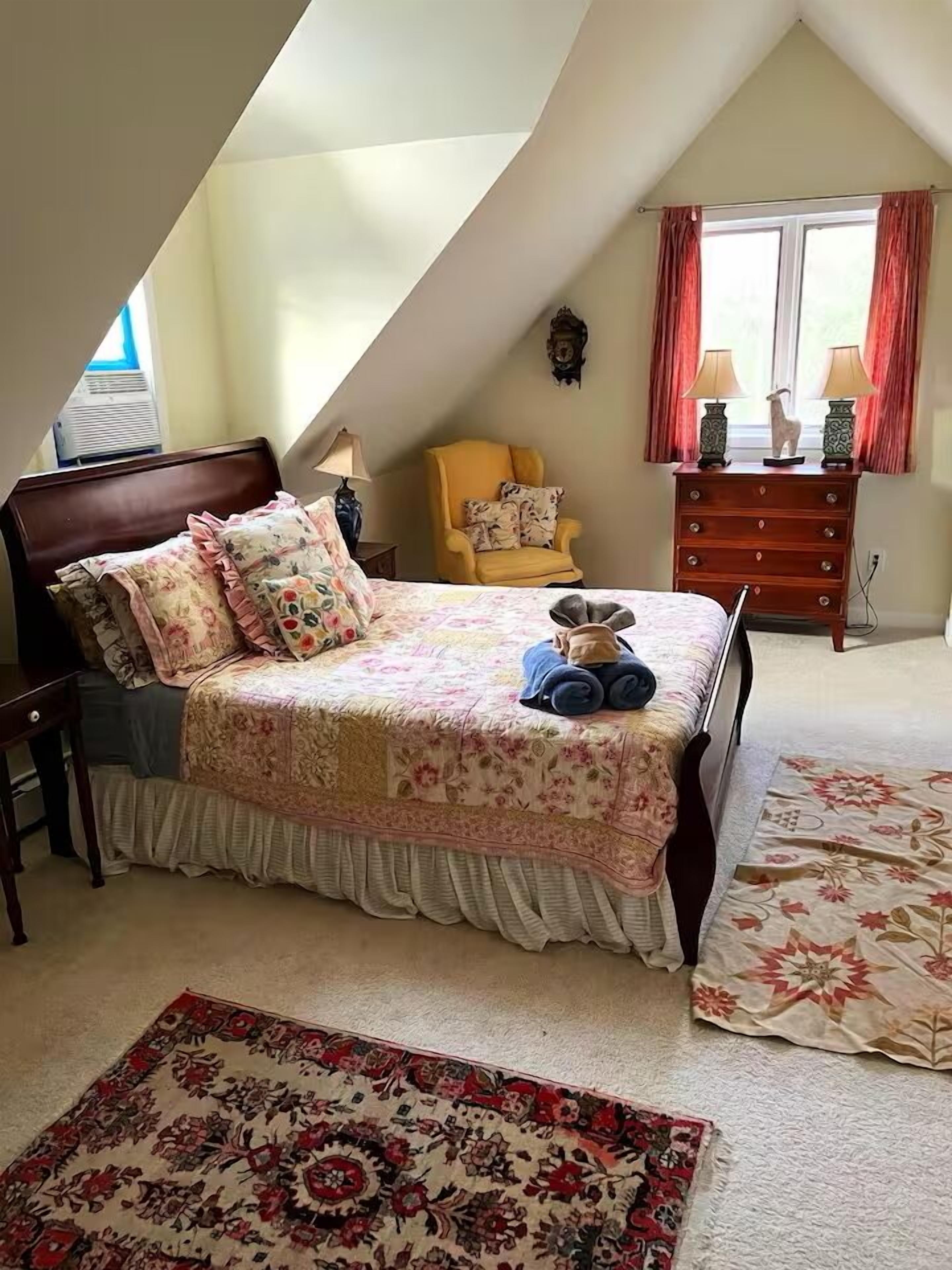 Un dormitorio con techo inclinado, una cama con ropa de cama floral, una silla, un aparador y dos lámparas.
