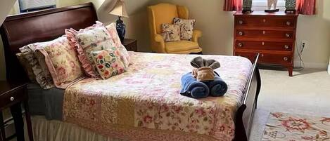 Un dormitorio con techo inclinado, una cama con ropa de cama floral, una silla, un aparador y dos lámparas.