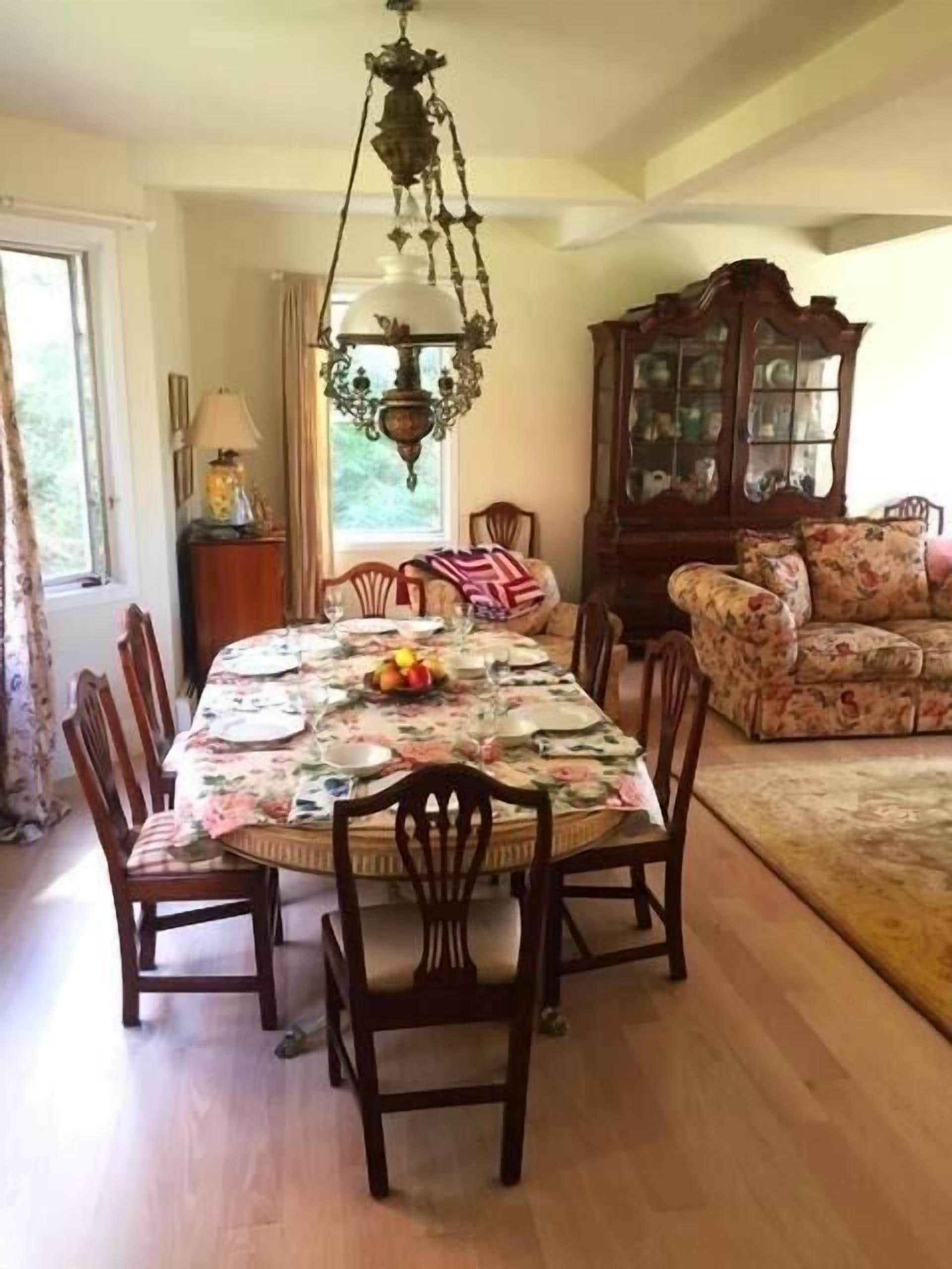 A dining area with a table set for four, surrounded by wooden chairs. A chandelier hangs above the table, and a floral-patterned sofa is visible in the background.