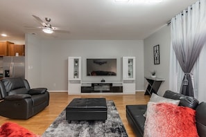 A modern living room with a flat-screen TV, black leather sofas, a black ottoman, and a ceiling fan. - Modern Townhome (Chapel Hill)