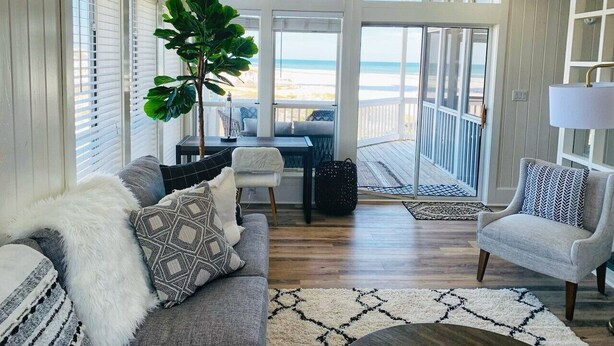 Uma sala de estar espaçosa, com uma grande janela com vista para a praia, um canto de estar acolhedor com sofá e poltronas, e uma mesa de jantar com cadeiras.