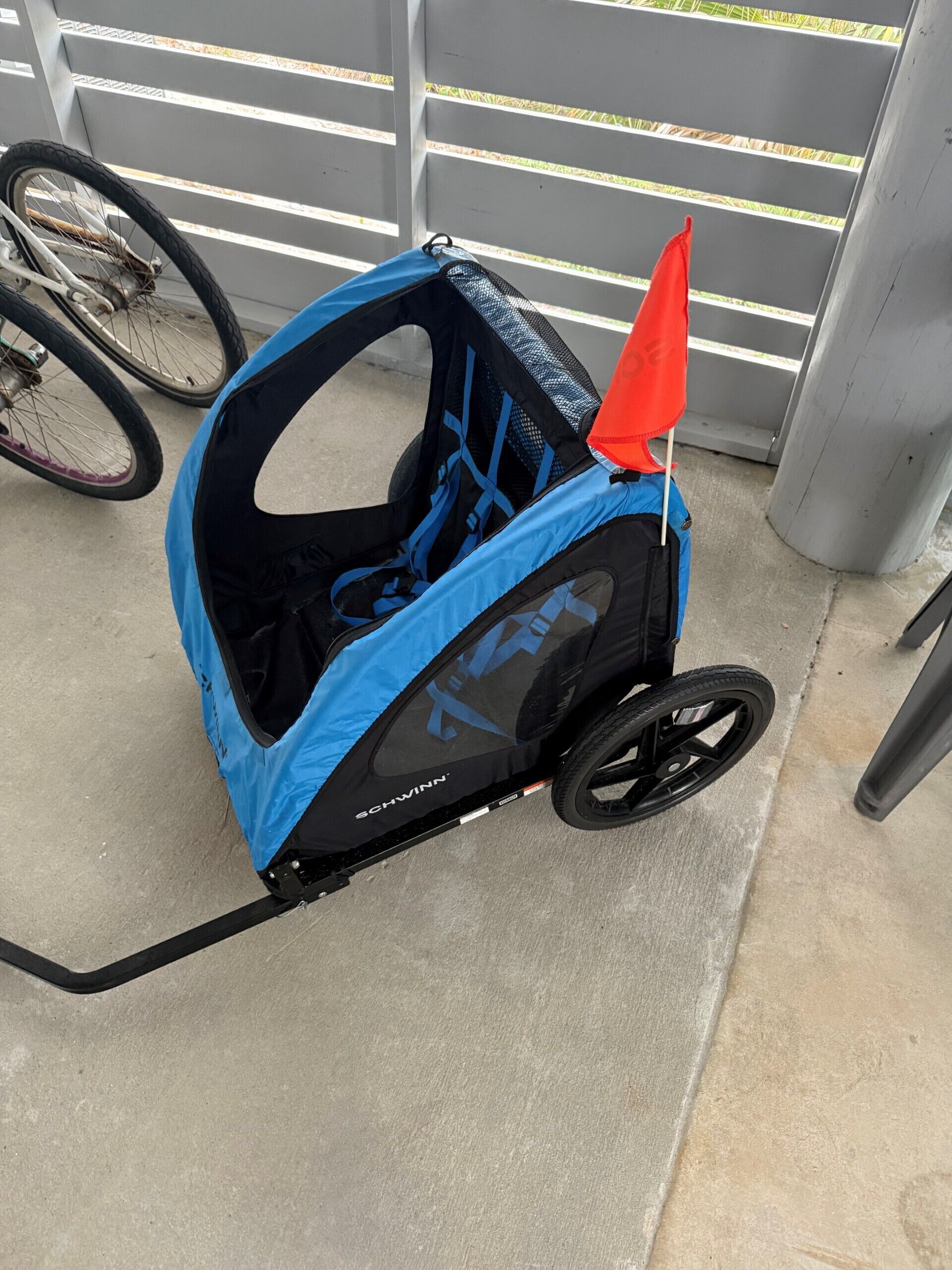 Une poussette pour bébé bleue et noire, surmontée d’un drapeau rouge, est garée sur une surface en béton.