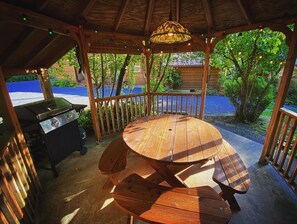 A wooden gazebo with a round table and benches, a grill, and string lights.