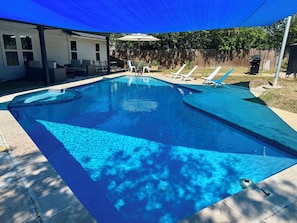 A rectangular swimming pool with a blue tint, surrounded by a concrete deck. There are lounge chairs and a covered patio area with a blue canopy.