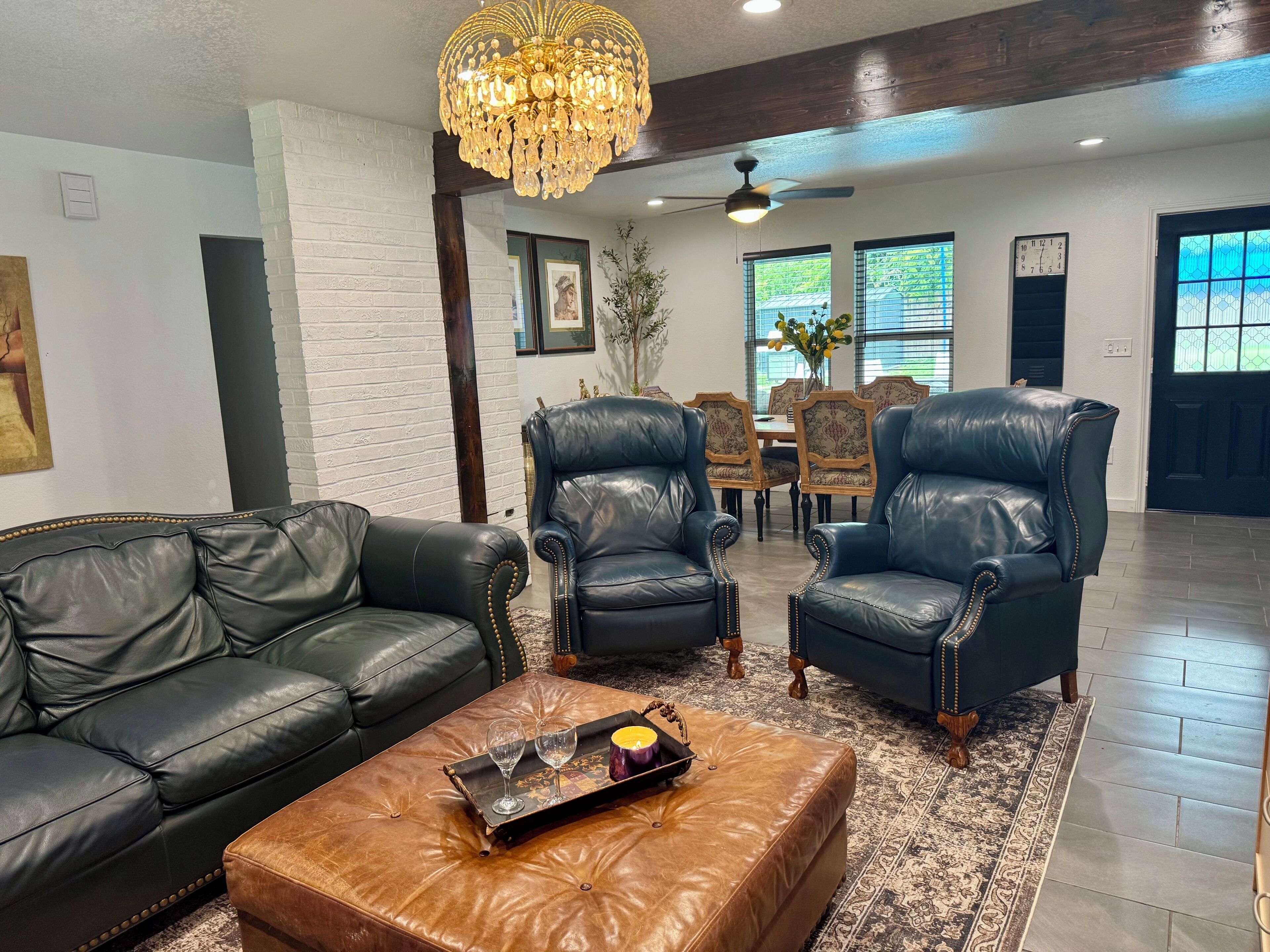 A living room with a black leather sofa, two leather armchairs, a brown ottoman, a chandelier, and a dining area in the background.