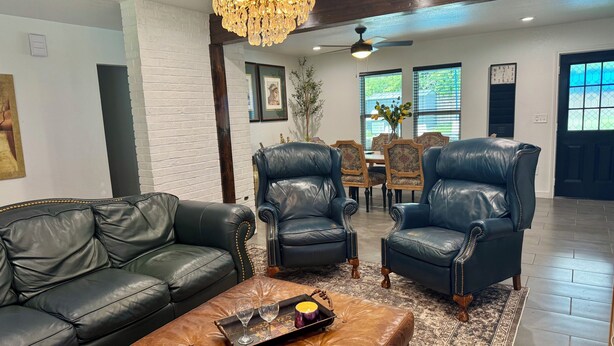 Un salon avec un canapé en cuir noir, deux fauteuils en cuir, un pouf marron, un lustre et, au fond, un coin repas.