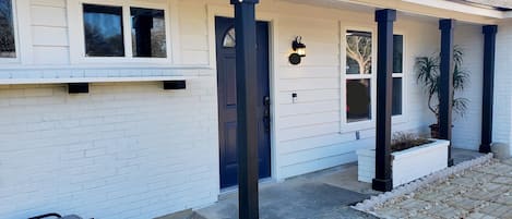 A two-story house with a covered porch, a front door, and a small garden area with a trash can and a water fountain.