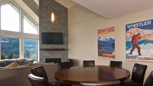 Una sala de estar moderna con una pared de acento de piedra, un televisor de pantalla plana, una chimenea y una mesa de comedor redonda con sillas. Hay dos pósteres en la pared, uno de Whistler, Canadá, y otro de Columbia Británica.