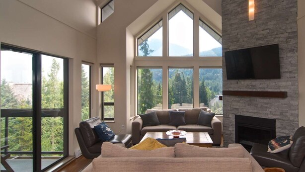 Amplia sala de estar con techo alto, ventanas grandes que ofrecen vistas al exterior, chimenea y televisor de pantalla plana montado en la pared.