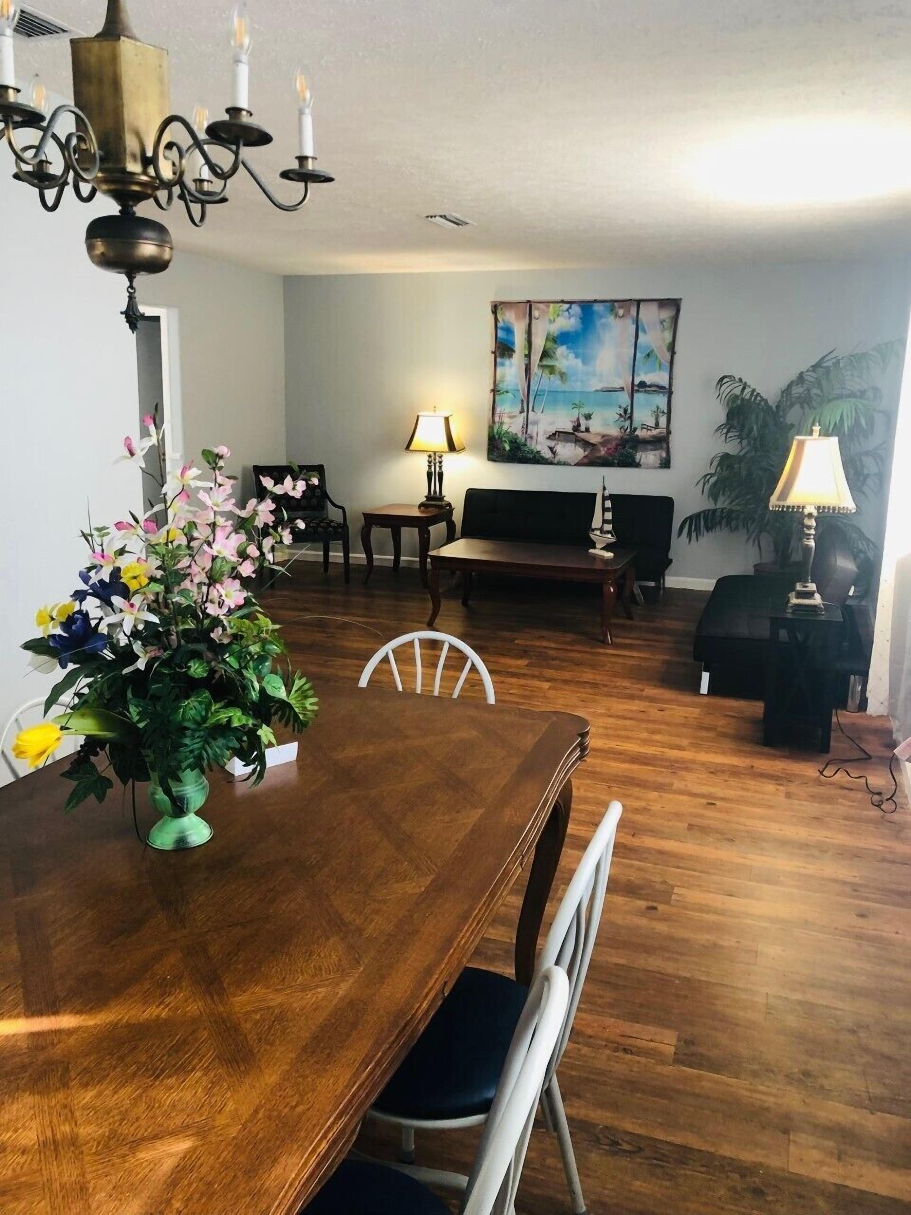 A dining area with a wooden table, chairs, a chandelier, a sofa, a painting, and a potted plant.