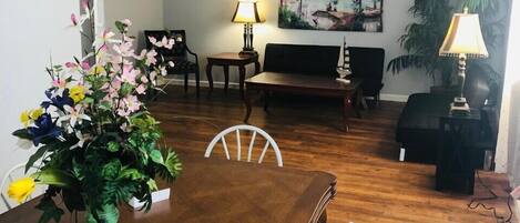 A dining area with a wooden table, chairs, a chandelier, a sofa, a painting, and a potted plant.