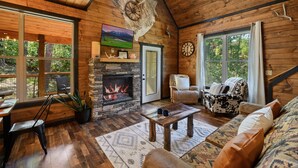 A cozy cabin interior with a stone fireplace, wooden walls, a leather sofa, and a dining area with a view of the outdoors. - Secluded! Game Room! Prkwy & Dollywood around the corner! Private deck! Hot Tub! (Sevierville)