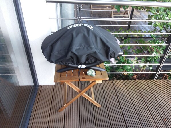 Une housse de barbecue noire recouvre un tabouret en bois sur un balcon doté d’une rambarde en métal et dont l’arrière-plan est constitué de feuillage verdoyant.