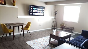 Ein Wohnzimmer mit Flachbildfernseher an der Wand, einem Esstisch mit zwei Stühlen, einer Couch, einem Couchtisch und einem gemusterten Teppich.