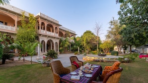 Un espacio al aire libre con área de comedor, una mesa dispuesta para una comida, rodeada de sillas de mimbre, frente a un edificio con arcos y un jardín.