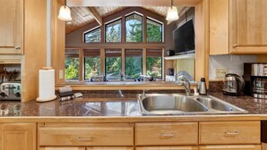 A kitchen with wooden cabinets, a granite countertop, a double sink, and a large window with a view of trees.