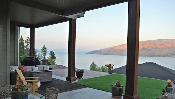 Eine überdachte Terrasse im Freien mit Blick auf einen See und Berge, ausgestattet mit Stühlen und einem Grill.