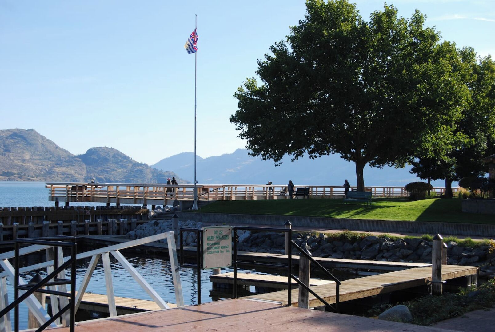 A waterfront park with a wooden dock, a flagpole with a flag, and a sign indicating the dock is closed from 9 am to 5 pm.