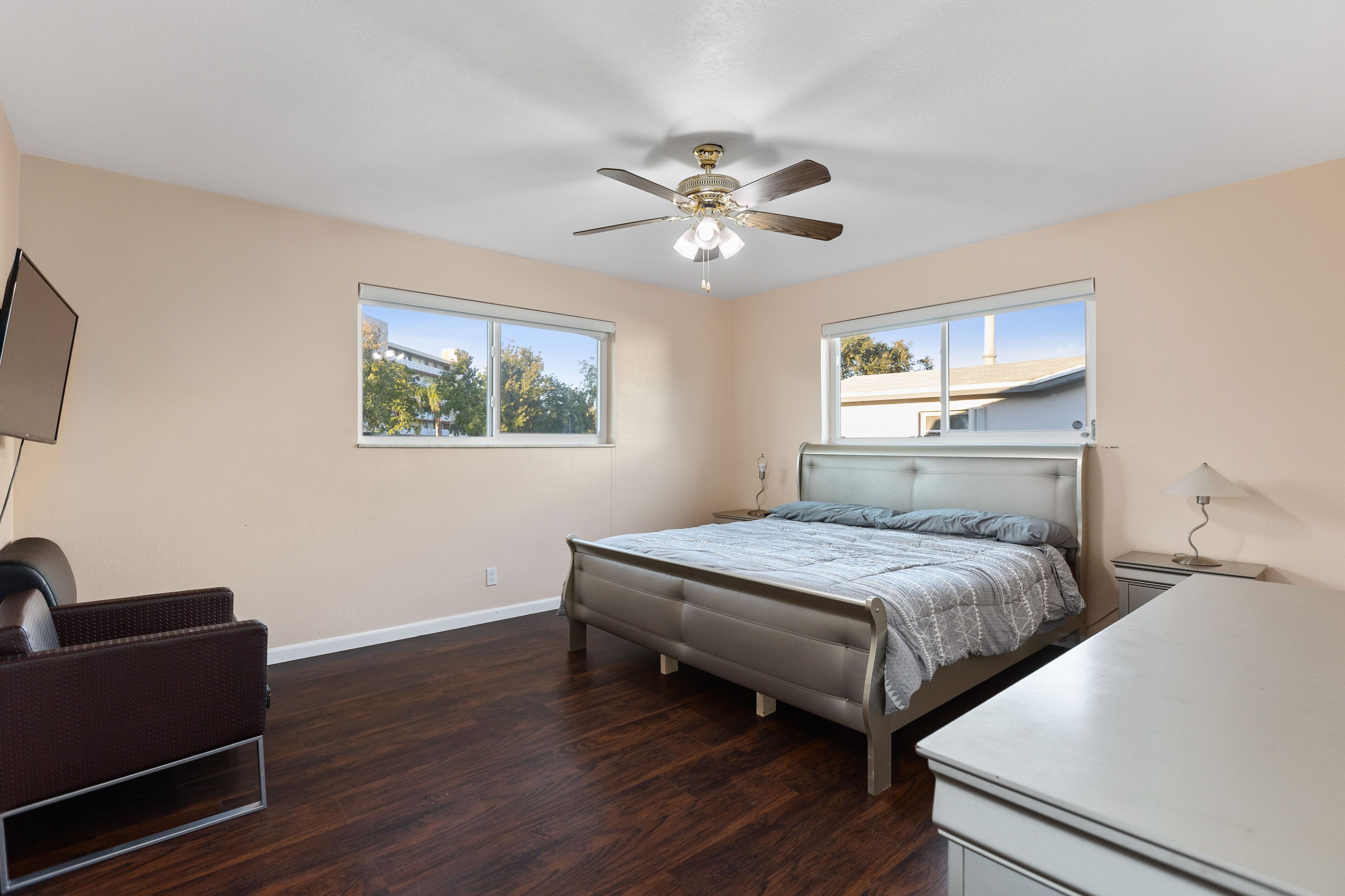 A bedroom with a bed, a desk, a chair, a ceiling fan, and two windows.