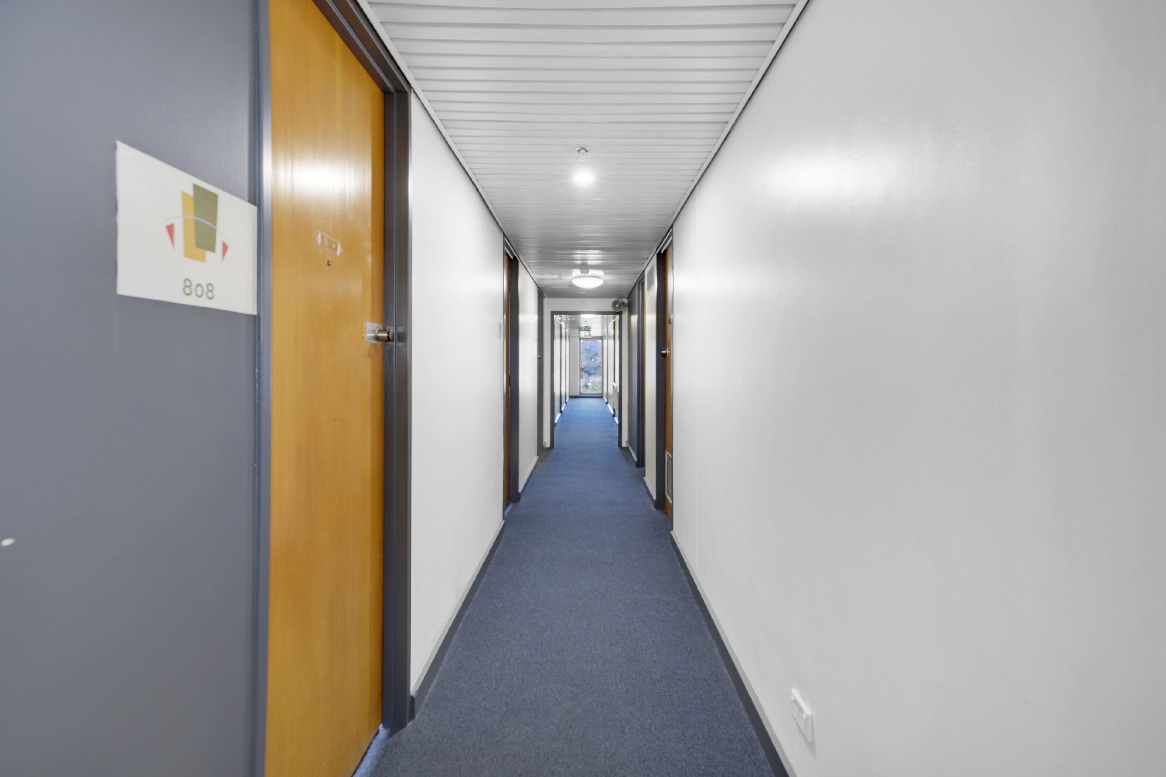 Un largo y estrecho pasillo con alfombra azul, puertas de madera a la izquierda y un letrero que indica la habitación 808.
