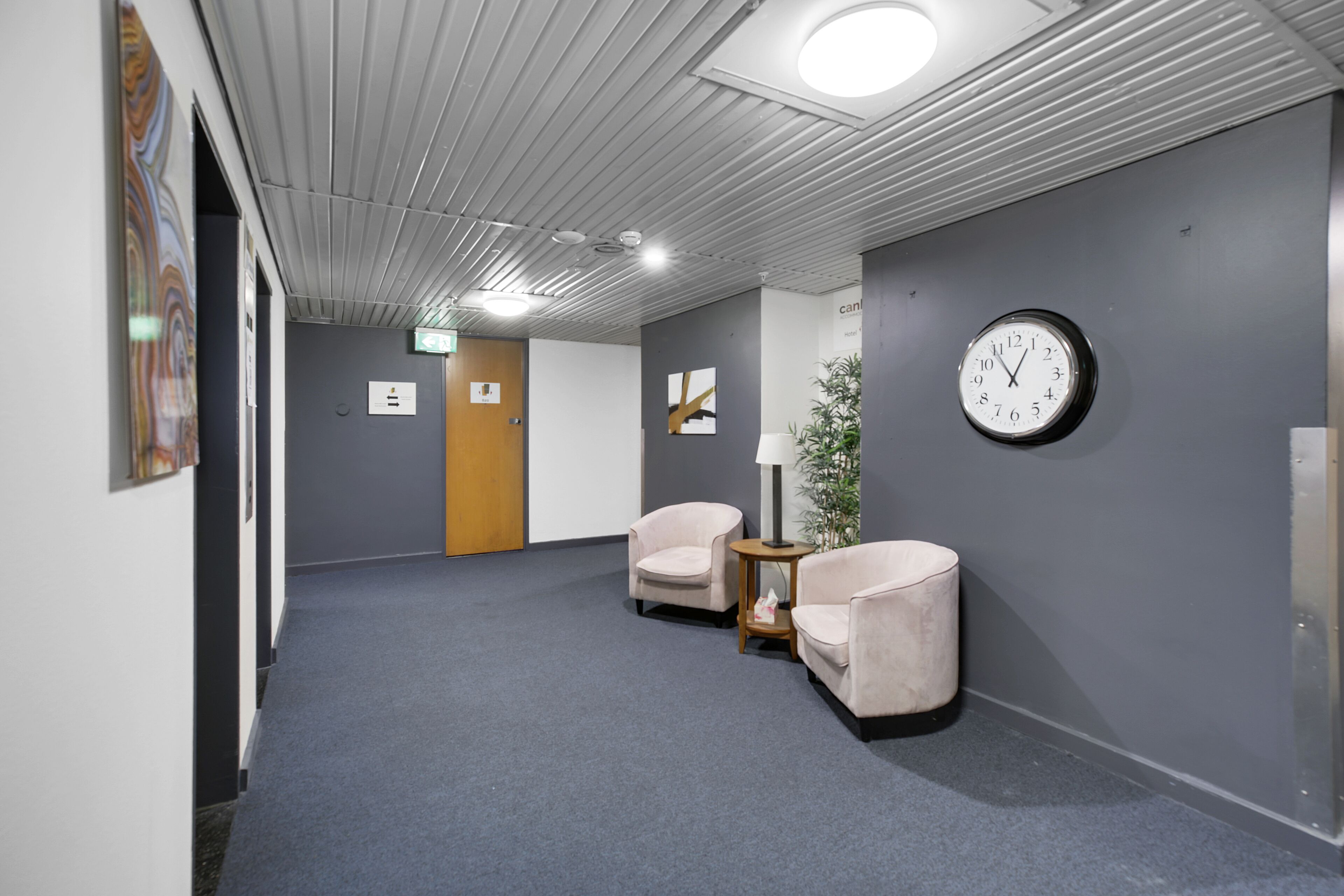 A hallway with grey walls, a clock, two chairs, and a small table with a lamp.