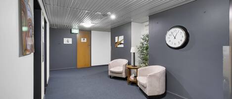 A hallway with grey walls, a clock, two chairs, and a small table with a lamp.