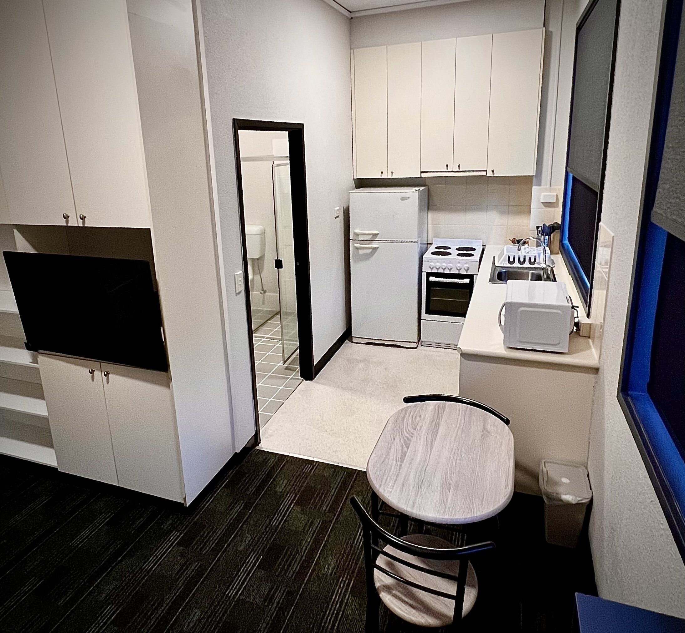 Cocina compacta con armarios blancos, un refrigerador, una estufa y un pequeño comedor con una silla.