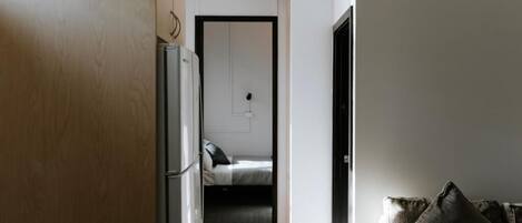 A hallway leading to a bedroom with a bed, a door, and a refrigerator.