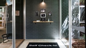 A self-check-in area with a black mat, a wall-mounted shelf with a QR code, and a sign offering assistance. - Canberra Accommodation Centre (Braddon)
