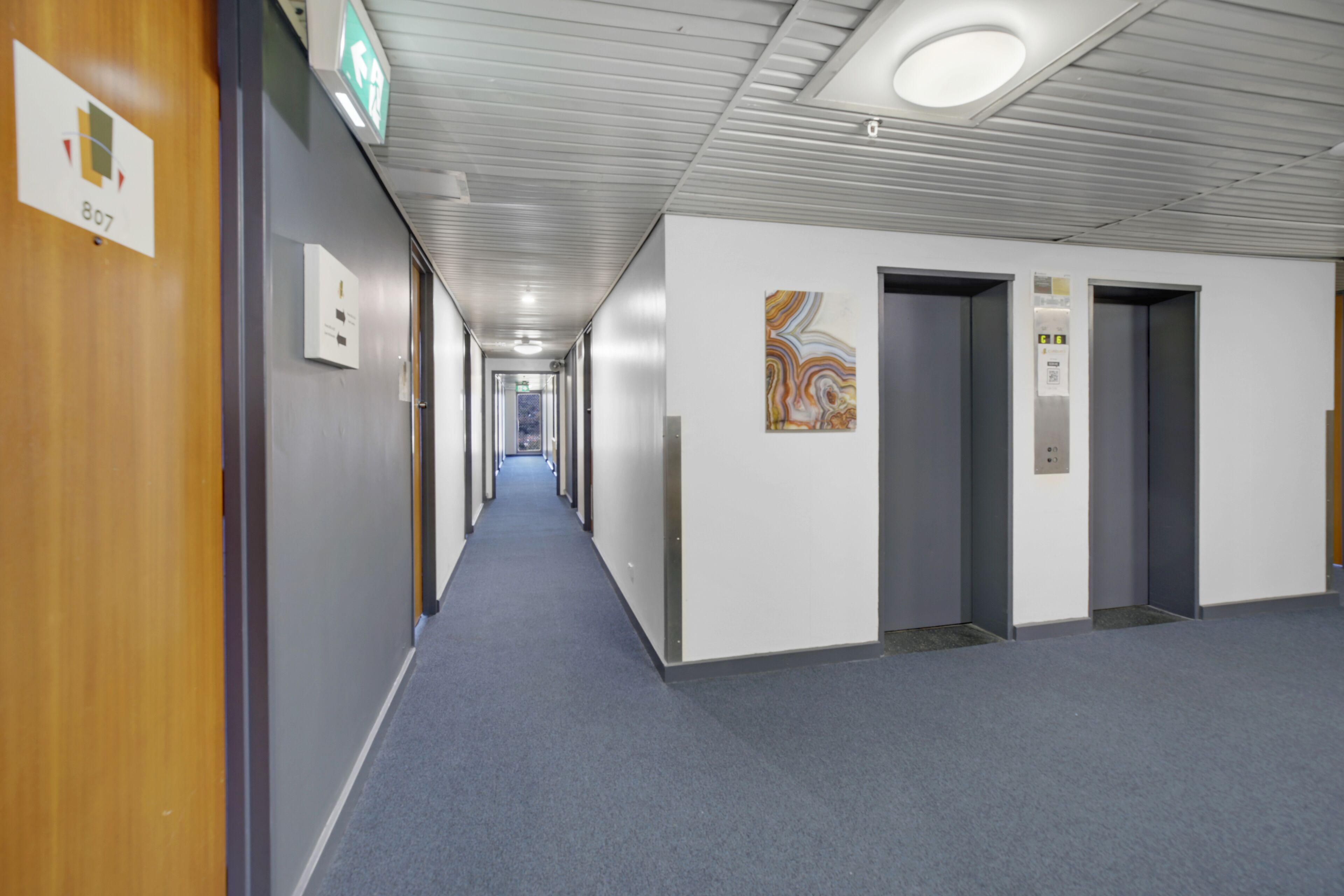 A long, narrow hallway with a blue carpet, wooden doors on the left, and a wall-mounted sign indicating an exit.