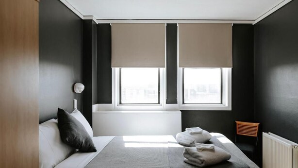 A modern bedroom with a bed, two windows with blinds, a radiator, and a small wooden chair. - Canberra Accommodation Centre (Braddon)