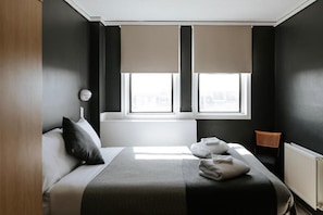 Un dormitorio moderno con una cama, dos ventanas con persianas, un radiador y una pequeña silla de madera.