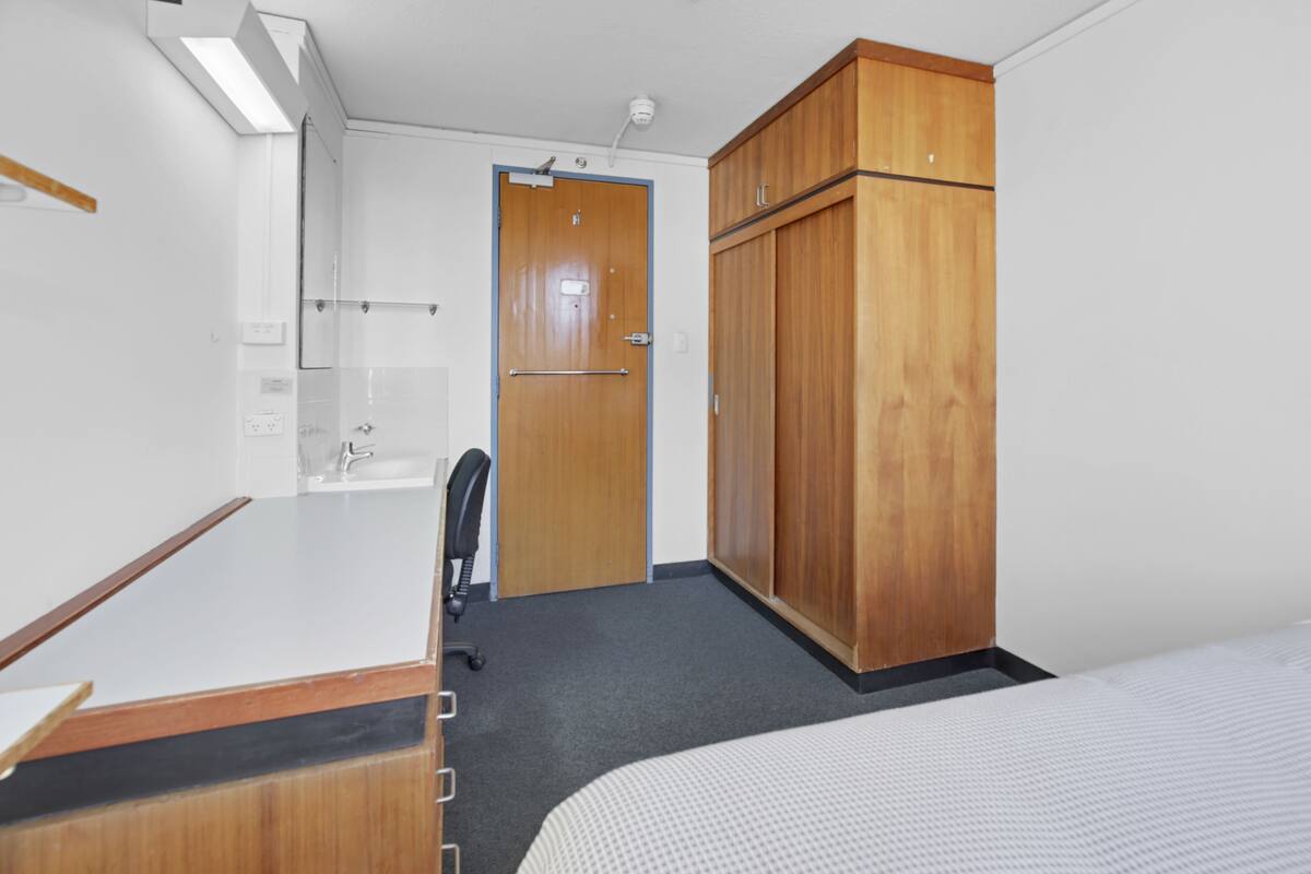 Ein Hotelzimmer mit Bett, Holzschrank, Schreibtisch mit Stuhl, Badezimmer mit Waschbecken und Spiegel sowie einer Tür zu einem weiteren Raum.