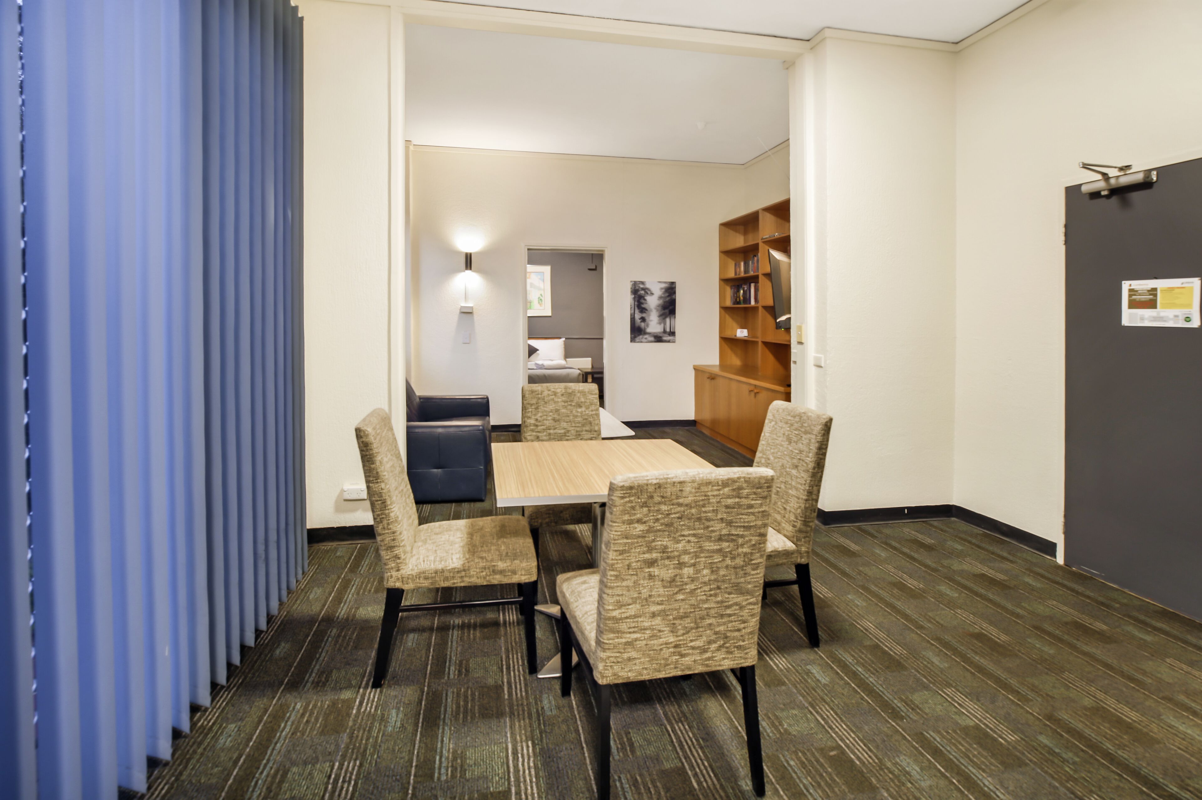 Habitación de hotel con comedor que incluye una mesa y cuatro sillas, una estantería, una pequeña cocina americana y una puerta con un cartel.