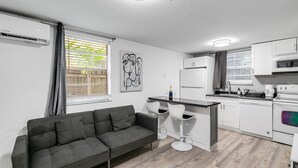Een moderne keuken met witte kasten, een zwart aanrecht en een ingebouwde oven. In de woonruimte staat een grijze bank en twee witte barkrukken.